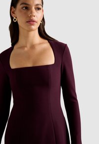 Langarmige jurk in diep bordeauxrood met een vierkante halslijn. Heeft een getailleerde pasvorm en een gladde stoftextuur. Gouden ringoorbellen.