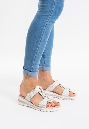 Person trägt hellblaue, hochgekrempelte Jeans und weiße Sandalen mit offenen Zehen und bedruckten Riemen vor weißem Hintergrund.