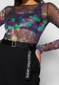 Transparante top met lange mouwen en multicolor abstracte patronen; gecombineerd met zwarte, high-waisted broek met een zwarte riem met logodetail.
