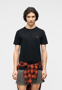 Νεαρός με κοντά σκούρα μαλλιά, που φοράει μαύρη μπλούζα με μικρό λογότυπο και κόκκινο-μαύρο πουκάμισο καρό δεμένο γύρω από γκρι σορτς.