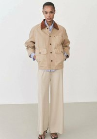 Beige canvas jacket with a brown corduroy collar, two front pockets, and button closure, paired with loose beige trousers.