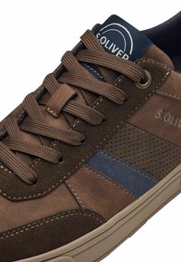 Basket décontractée marron avec une tige en suède et cuir texturé, présentant une bande d'accent bleue, des panneaux de lacets à motifs et une languette de marque.