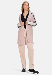 Knielanger, hellrosa Cardigan mit gerippter Textur, offener Vorderseite und kontrastierenden gestreiften Ärmeln. Über einem schwarzen Oberteil und hellen Hosen getragen.