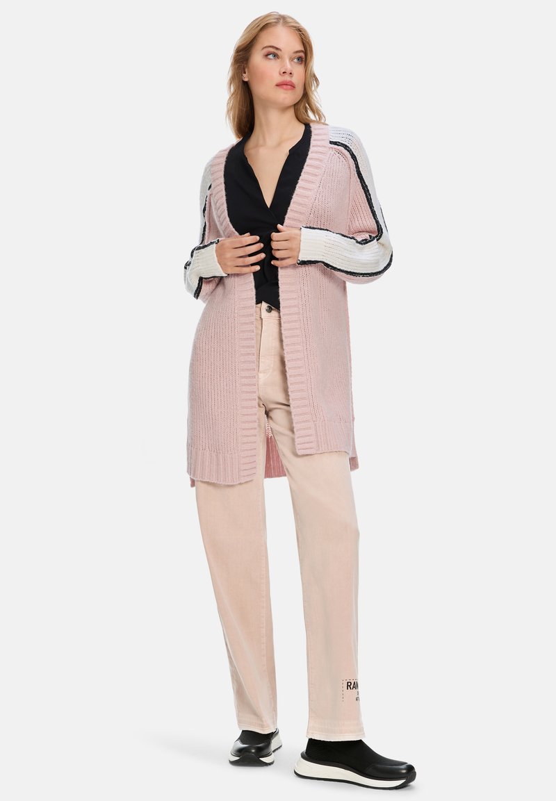 Knielanger, hellrosa Cardigan mit gerippter Textur, offener Vorderseite und kontrastierenden gestreiften Ärmeln. Über einem schwarzen Oberteil und hellen Hosen getragen.