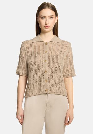 Femme portant un cardigan beige à manches courtes, en maille ajourée avec des boutons en bois, et un pantalon beige, debout devant un fond uni.