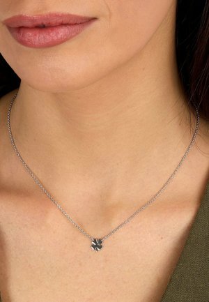 Nærbillede af en kvindes hals, der bærer et delikat sølvkæde med en lille firekløver vedhæng.