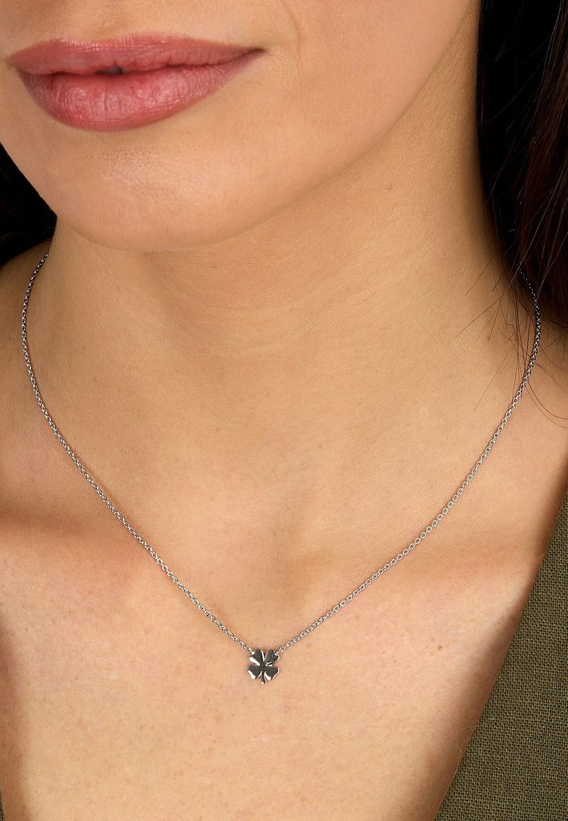 Gros plan sur le cou d'une femme portant un collier délicat en argent avec un petit pendentif en forme de trèfle à quatre feuilles.