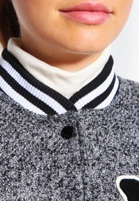 Grå tekstureret jakke med en sort og hvid stribet krave, der har en sort knaplukning og broderet detaljer på brystet.