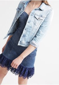 Jasnoniebieska kurtka dżinsowa z zapięciem na guziki, dwoma kieszeniami na piersi i podwiniętymi rękawami, zestawiona z gorsetową sukienką dżinsową w kolorze granatowym, ozdobioną koronkowym wykończeniem.