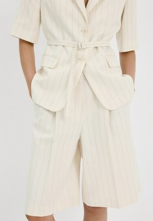 Traje beige de rayas con mangas cortas y pantalones cortos a juego hasta la rodilla, con cintura ceñida con cinturón y bolsillos con solapas.