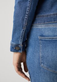 Blauwe denimjas mouw met knoopsluiting, met zichtbare textuur en stikseldetails, gecombineerd met blauwe denimjeans met achterzak.
