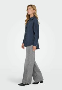 Donna in piedi di lato che indossa una camicia blu navy larga con bottoni, jeans grigi a gamba larga e stivali neri con tacco su uno sfondo chiaro uniforme.