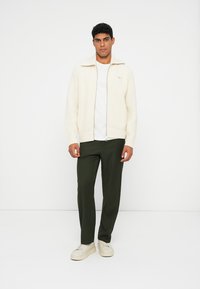 Homem em um suéter branco de zíper canelado, camiseta branca, calças verde-escuras e tênis brancos, apoiado contra um fundo branco liso.