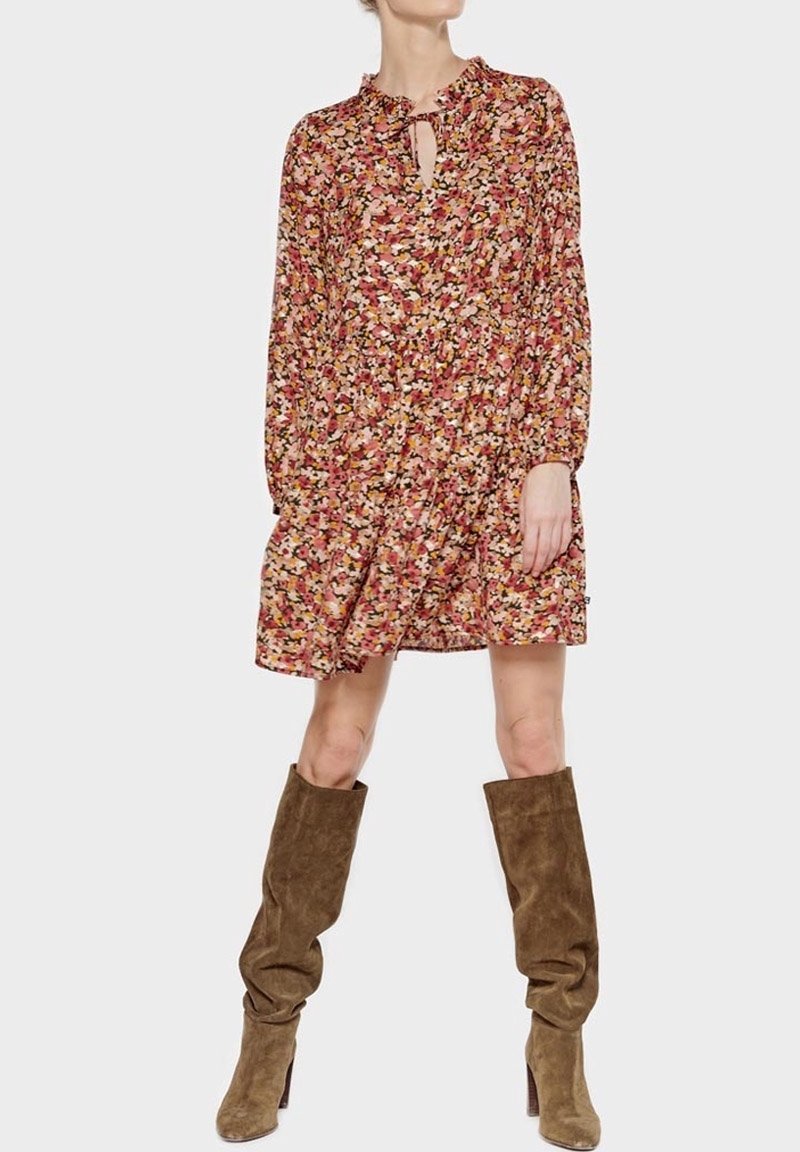 Femme portant une robe longue à manches longues à motif floral avec détails à volants, associée à des bottes hautes en daim marron décontractées, sur un fond uni.