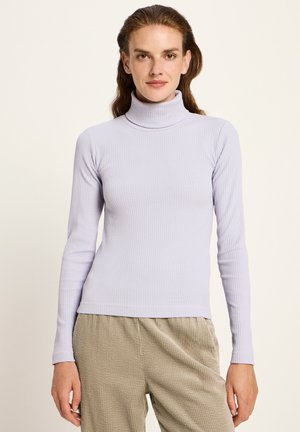 Frau, die einen helllila gerippten Rollkragenpullover und beigefarbene, strukturierte Hosen trägt, und vor einem einfarbigen hellen Hintergrund steht.