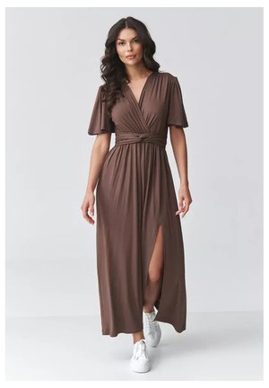 Kobieta idąca do przodu w brązowej sukience kopertowej z krótkimi rękawami i przednim rozcięciem, w połączeniu z białymi trampkami, na jednolitym tle.