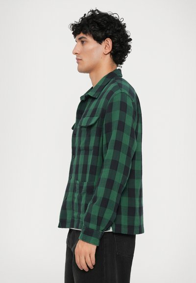 Camisa de flanela xadrez verde e preta com fecho de botões à frente, dois bolsos no peito e mangas longas. Textura suave e corte descontraído.
