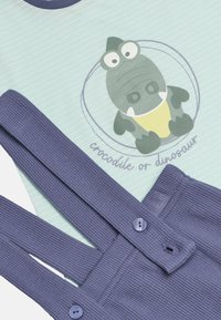 Top de algodón con rayas menta, que presenta un gráfico de un cocodrilo/dinosaurio de dibujos animados, combinado con un peto de canalé azul marino con cierres de botón.