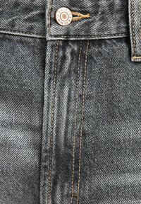 Lähikuva mustista denim-farkuista, joissa on beige ompelu ja metallinappi, jossa lukee "Bershka Denim."