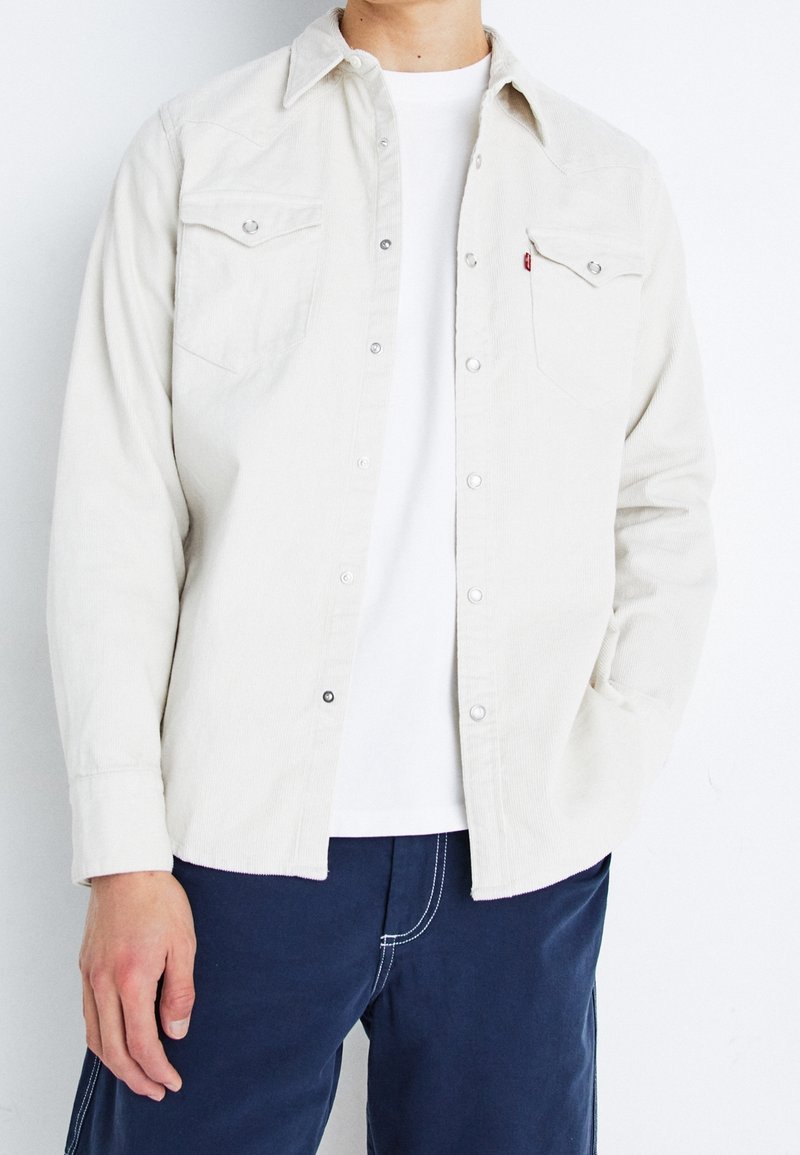 Homme portant une chemise en velours côtelé blanche boutonnée sur un t-shirt blanc, associée à un pantalon bleu foncé avec des coutures blanches.