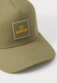 Olivengrønn cap med en strukturert form, mesh bak og et firkantet merke med "Brixton"-logoen i gult broderi. Teksturert fabrikkdetalj.