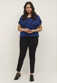 Blåblommig topp med korta ärmar, kombinerad med svarta slim-fit byxor och svarta slip-on skor, mot en neutral bakgrund.