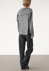 Persona con capelli biondi corti che indossa un top grigio a maniche lunghe con trama e jeans neri larghi mentre cammina su un pavimento semplice contro un muro bianco.