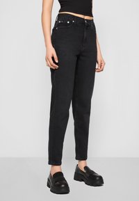 Höga svarta jeans med en figurnära silhuett, femficksdesign, metallbeslag och lätt blekningar på tyget. Modell som bär svarta skor.