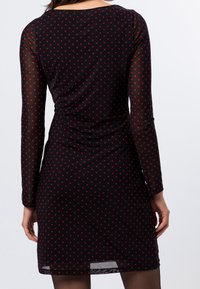 Femme portant une robe noire ajustée avec de longues manches transparentes et des pois rouges, vue de dos sur un fond blanc uni.