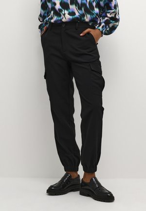 Osoba nosząca czarne spodnie dresowe typu cargo z elastycznymi ściągaczami i czarne skórzane loafersy, ręce w kieszeniach oraz niebiesko-zieloną wzorzystą bluzkę.