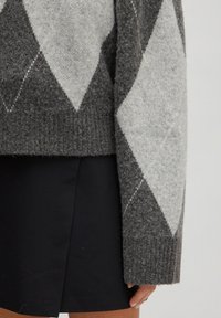 Manche de pull à losanges gris et gris clair sur une jupe noire, montrant une main reposant à côté de l'ourlet de la jupe.