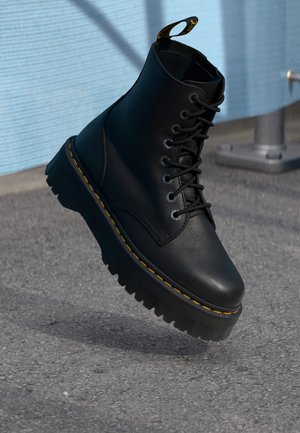 Zwarte leren veterschoen met dikke geribbelde zool en gele stiksels, hangend boven asfalt nabij een blauw metalen hek.