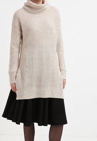 Kobieta w kremowym swetrze z grubej dzianiny z golfem, nosząca czarną spódnicę w kształcie litery A i przezroczyste czarne rajstopy, stojąca na tle białej ściany.