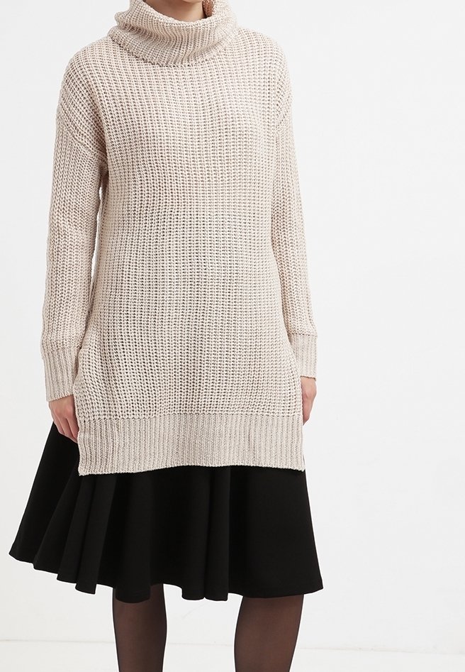 Kobieta w kremowym swetrze z grubej dzianiny z golfem, nosząca czarną spódnicę w kształcie litery A i przezroczyste czarne rajstopy, stojąca na tle białej ściany.