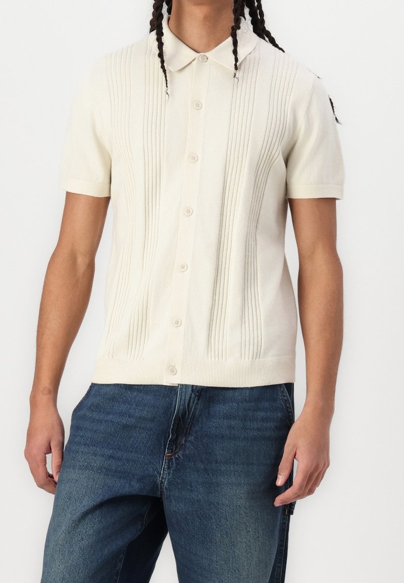 Personne portant une chemise crème en maille à manches courtes avec des boutons et un détail de côtes verticales, associée à un jean en denim bleu.