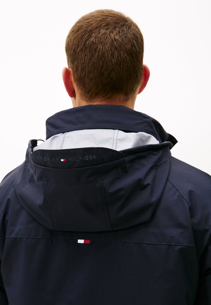 Marinblå huva jacka med grå innerfoder. Har en logotyp broderad på kragen och en liten tri-färgad accent nära baksidan av fållen. Slät textur.