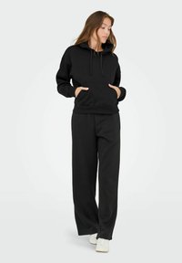 Sort hættebluse med frontlomme og snore, parret med løstsiddende sorte sweatpants og hvide sneakers. Blød stoftekstur.
