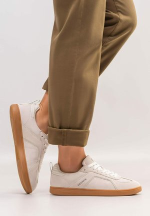 Person trägt beige Hose mit aufgekrempelten Säumen und weiße Low-Top-Sneaker mit Gummisohlen, steht mit einem leicht angehobenen Fuß.
