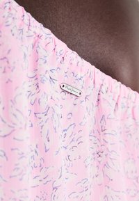 Gaiši rozā blūze ar ziedu rakstu, elastīgu apkakli un metāla birku ar uzrakstu "Pepe Jeans." Audums izskatās teksturēts un viegls.
