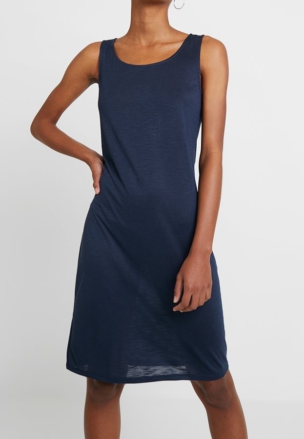 Personne portant une robe sans manches bleu marine arrivant aux genoux, debout avec une main sur la hanche sur un fond uni.