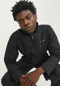 Zwarte zip-up jas van gladde, lichtgewicht stof. Heeft eenstaand kraag en een klein logo detail op de borst. Aangepaste pasvorm.