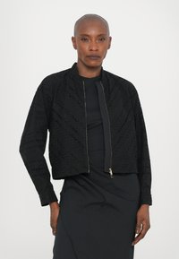 Čierna bomber bunda s komplexným detailným výrezom, zipsom uzatvárania a skráteným strihom. Obsahuje dlhé rukávy a texturovaný materiál.