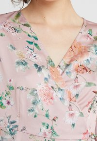 Blouse fleurie en tissu rose doux avec un col en V, présentant un motif aquarelle de fleurs pêche et blanches avec des feuilles vertes.