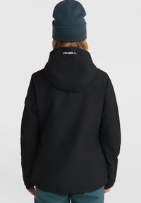 Schwarze Jacke aus weichem, strapazierfähigem Stoff mit Kapuze und Logo auf dem Rücken. Die Person trägt eine dunkelblaue Mütze.