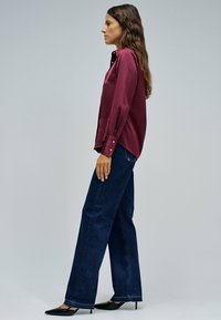 Camisa de satén burdeos con botones y mangas largas, combinada con jeans de pata ancha azul oscuro y tacones negros de punta.