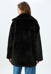 Sort faux fur frakke med oversized krave, afslappet pasform, tekstureret overflade og sænkede skuldre, båret over blå jeans.