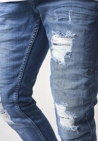 Blue denim jeans featuring a slim fit with distressed patches and frayed edges. The fabric displays a typical denim texture.