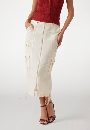 Vrouw draagt een beige midi-rok met rits aan de voorkant en cargozakken, gecombineerd met bruine sandalen met bandjes en een rood mouwloos topje.
