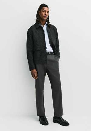 Bărbat cu părul împletit, purtând o jachetă neagră cu pete, cămașă bleu, pantaloni gri și pantofi negri, stând în fața unui fundal alb.