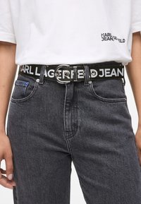 Cinturón trenzado negro con una hebilla circular de plata, que presenta el texto grabado "KARL LAGERFELD JEANS" en letras blancas. Usado con jeans grises.
